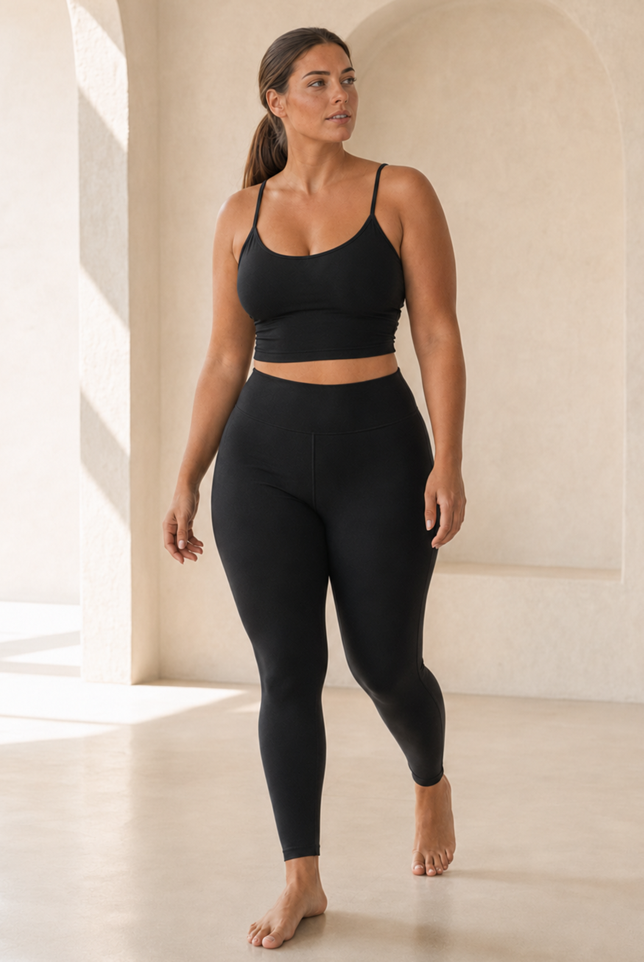 A woman modeling a black activewear set consisting of a spaghetti-strap crop top and high-waisted leggings, standing in a minimalist, sunlit room with beige walls and floor.
