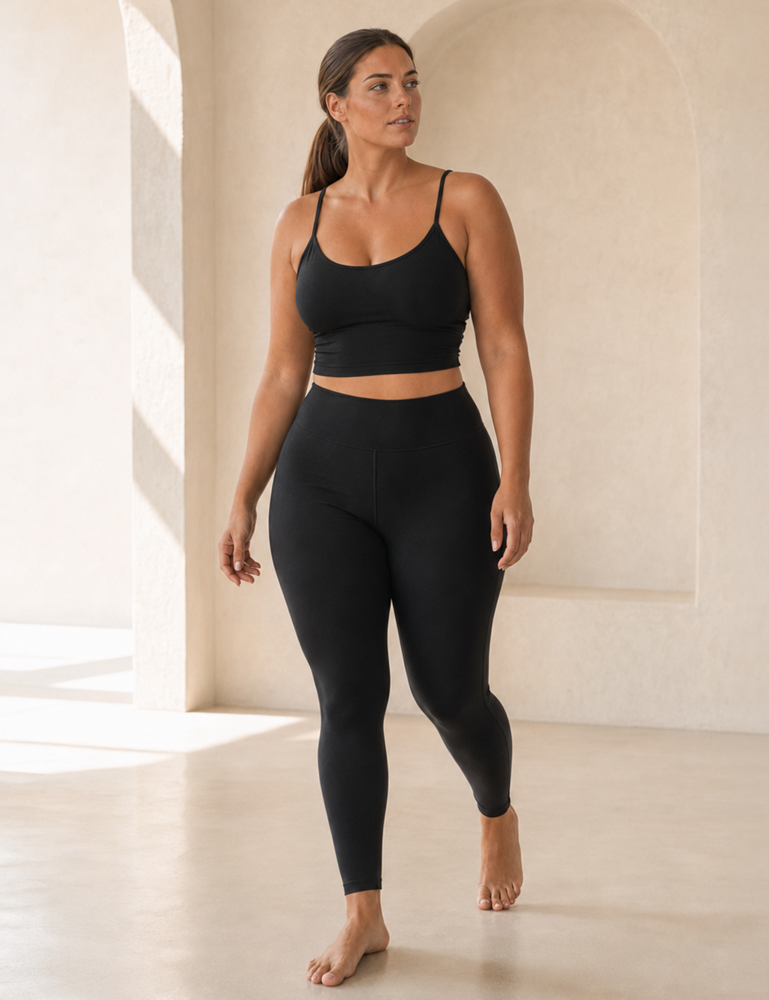 A woman modeling a black activewear set consisting of a spaghetti-strap crop top and high-waisted leggings, standing in a minimalist, sunlit room with beige walls and floor.